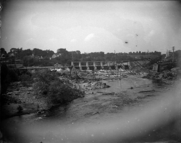 Elevated view of a dam on a river. There is a collapsed bridge in front of the dam, and people standing on the road on the right. Identified as possibly the Black River Falls.