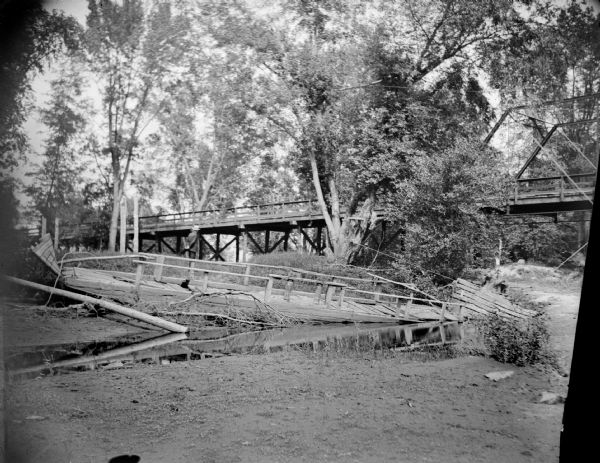 View towards section of a collapsed bridge along a riverbank. There is another bridge spanning the river in the background.
