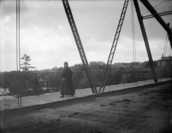 View across bridge towards a Native American woman walking on the wooden sidewalk of a bridge above a river. Identified as the Harrison Street bridge. Buildings are in the background.