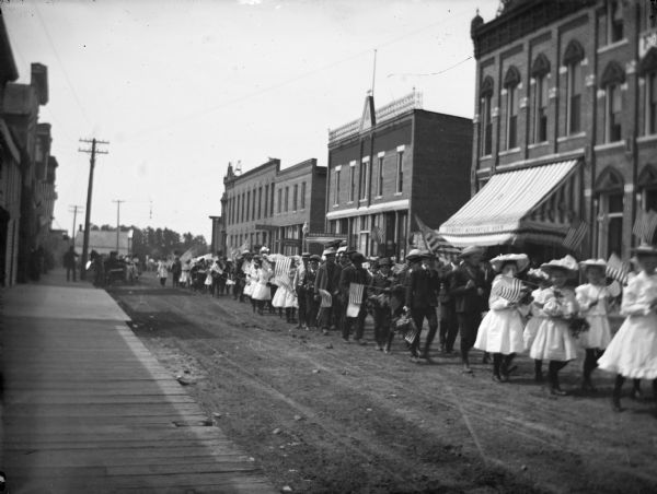 View down wooden sidewalk and unpaved street towards a long line of children parading up the street. Many of the children are carrying American flags. Identified as a Memorial Day celebration with the location identified as Water Street looking north between Main and Harrison Streets.