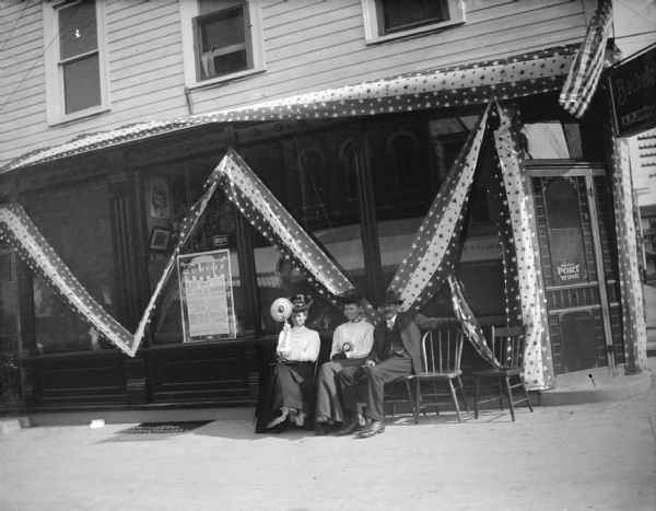 View from street towards a man and two women sitting in front of a storefront decorated with patriotic streamers. The woman on the left is holding a hand fan. Identified as the A.P. Johnson Tavern on Independence Day.