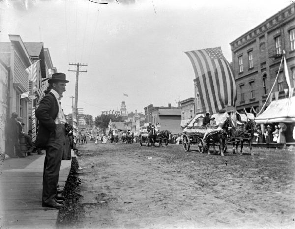 View down sidewalk towards people standing along a street in town and watching a parade. Coming down the unpaved street are people on decorated horse-drawn wagons. A large U.S. flag is suspended over the street. Identified as a parade on Main Street.