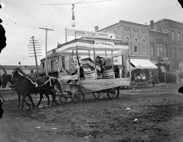 View towards a man posing sitting in a decorated wagon pulled by a team of two decorated horses on a town street. Banners on the wagon read: "The Old Standby" and "Badger State Banner, 1856-1905."