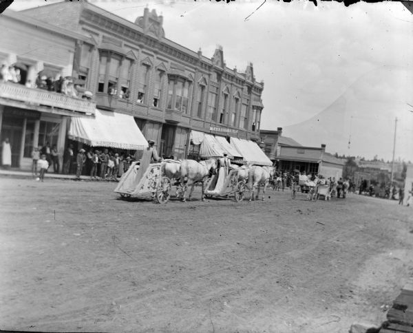 View down street towards of women posing standing in chariots pulled by teams of two horses. They are moving down the street in front of wooden sidewalks lined with people. A crowd of people are standing on a balcony on the far left.