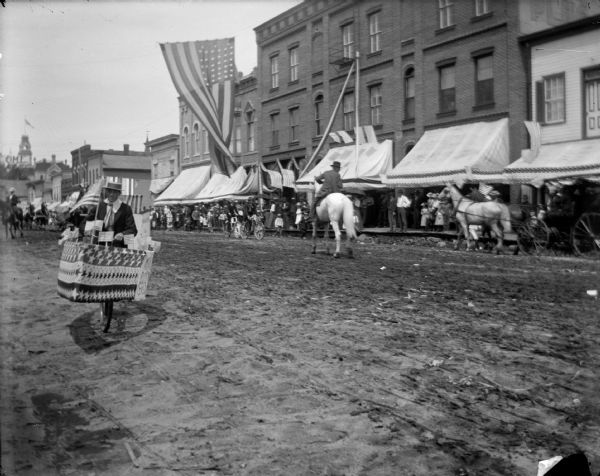 View across unpaved town street during a parade. There is a man in the left foreground wearing a costume that includes a U.S. flag, and a decorated bicycle with banners and signs. A large U.S. flag is suspended over the street, and crowds are watching from the opposite side of the street in front of storefronts.