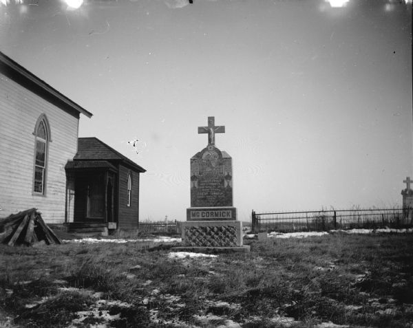 View towards a tombstone in the side yard of a church. There are patches of snow on the ground, and a fence and another tombstone in the background. Patches of snow are on the ground. Identified as the memorial for Christopher and Ann McCormick.