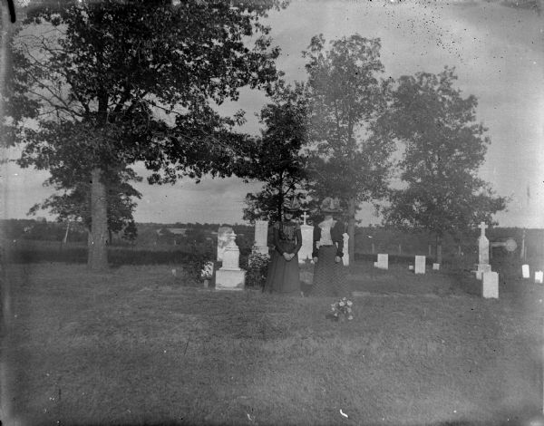 View across grass towards two women posing standing in front of a group of tombstones in a cemetery.