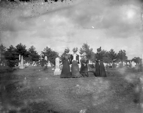 View across grass towards three women and one girl posing standing in a cemetery. The woman on the right is holding a closed umbrella.