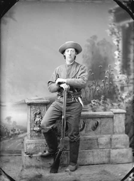Full-length studio portrait of a man standing with his foot on a prop stone wall in front of a painted backdrop. The man is posing standing and leaning on a rifle, and is wearing a belt with bullets and a knife, a hat with an upturned brim, and leggings over his trousers.