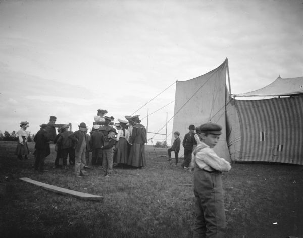 View across grass towards men, women, and children standing outside a tent, possibly a Chautauqua tent. A boy wearing a cap is standing with his arms crossed in the foreground.