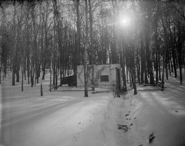 View down snow shoveled path towards a hunting shack in a forest surrounded by a wood fence.
