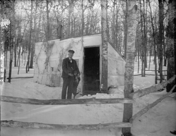 View across fence towards a man posing standing holding a dog up on its hind legs in front of a hunting shack in a forest in deep snow.