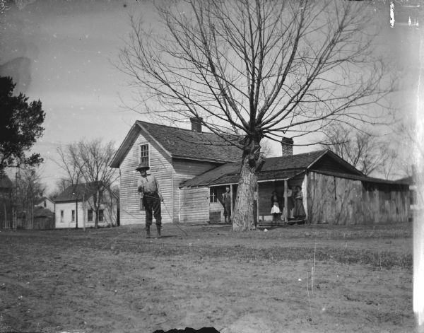 View across yard towards a man standing and twirling a lasso rope in front of a wooden house. Three women and a man are standing and watching from the porch.