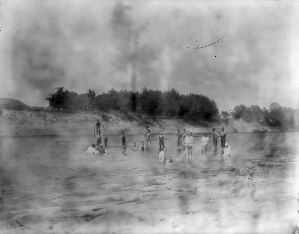 View towards a group of men, women, and children wading in shallow water. The far shoreline has a steep bank, with a fence and trees.
