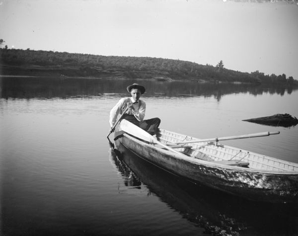 A man is sitting in the back of a canoe or rowboat holding a paddle in the water by his side. The canoe or rowboat also has oars in oarlocks. On the far shoreline are trees on a ridge.