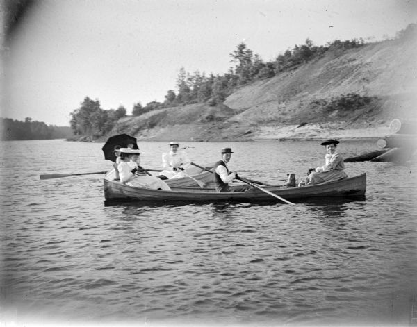 View across water towards four women and two men posing sitting in two rowboats on the water, with a steep shoreline in the background.