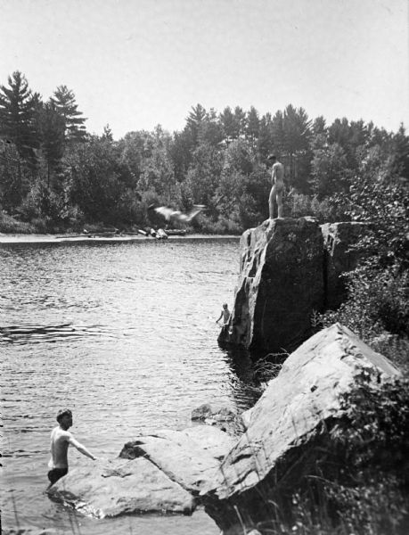 Copy photograph of an elevated view of a nude man standing on a rock bluff along a river, two men standing in the water below, and another man in mid-air jumping into the river. Identified as the swimming hole in the Black River just south of the cemetery at Spaulding Rock before the damming of the river. 