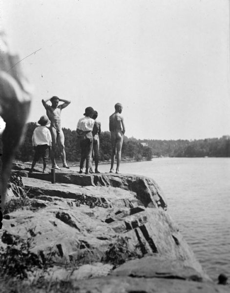 Copy photograph of an outdoor view of two nude men and three boys standing on a rock formation above a river. Identified as the swimming hole in the Black River just south of the cemetery at Spaulding Rock before the damming of the river.