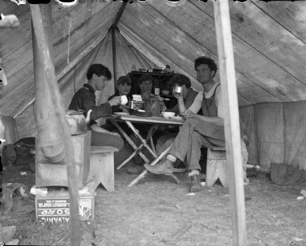Group portrait of five men sitting around a table inside a tent eating.