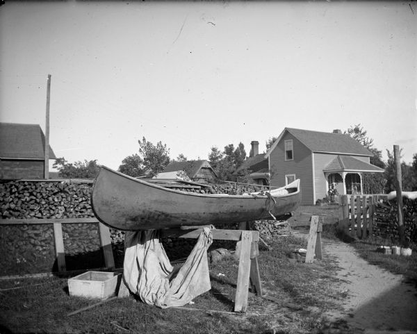 A canoe on wooden sawhorses is near a stack of firewood near a fence. In the background is a house and outbuildings in a yard.