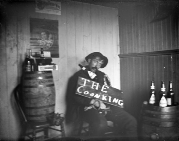 Portrait of a man sitting indoors in a chair with his eyes closed. A bottle is resting on his shoulder, and a sign has been placed on his lap that reads: "The Coon King." Bottles are sitting on top of barrels nearby.