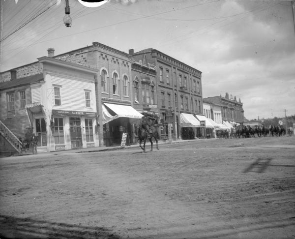 View across intersection towards a boy on horseback. Further down the unpaved street lined with storefronts is a large group of horses coming towards the intersection. Location identified as probably Main Street in Black River Falls looking east.