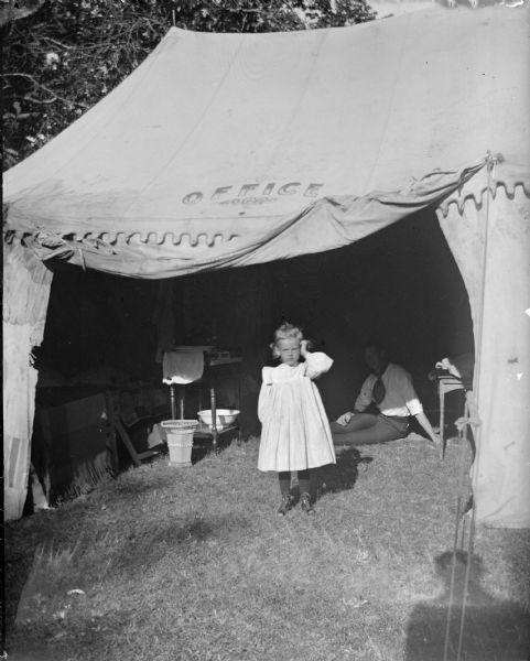 View towards a girl standing in the open doorway of a tent. In the background is a man sitting on the grass inside the tent. Probably the Chautauqua Office. A sign stenciled above the tent door reads: "Office."