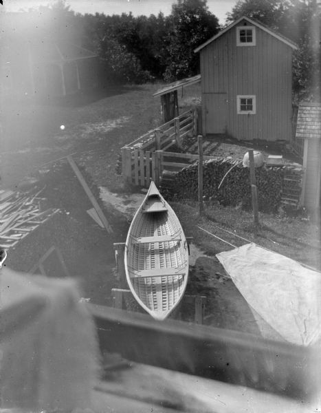 View from above, from a roof or balcony, of a canoe on sawhorses near firewood stacked in a yard near a fence. Two buildings are in the yard.