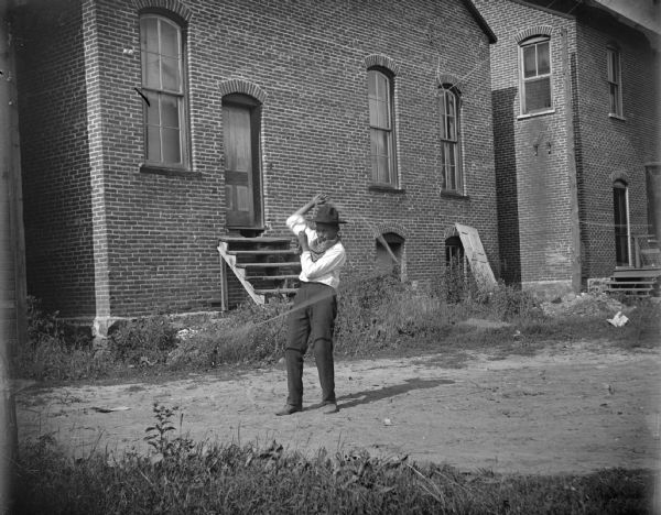 View towards a man standing and twirling a lasso in the dirt yard of a large brick building. The building is identified as the Opera House.