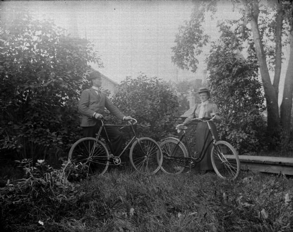 Outdoor portrait of a man and a woman posing standing in the grass and displaying bicycles in front of bushes and trees. In the background is a building.
