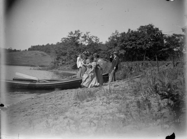 View along sandy shoreline towards three women and a man standing near a rowboat pulled up onto the shore. One of the women is holding an infant in her arms. The man is looking through a pair of binoculars.