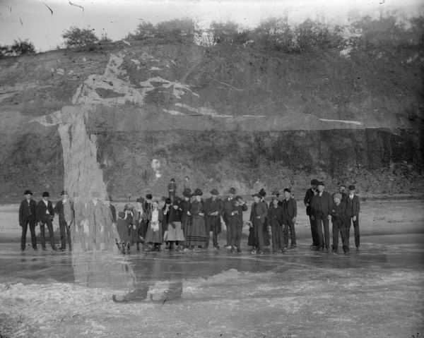 View across ice towards a large skating party posing for a group portrait. In the background along the shoreline is an exposed cliff with bushes and trees along the top. The image also includes a double exposure of a man standing on ice and wearing ice skates.