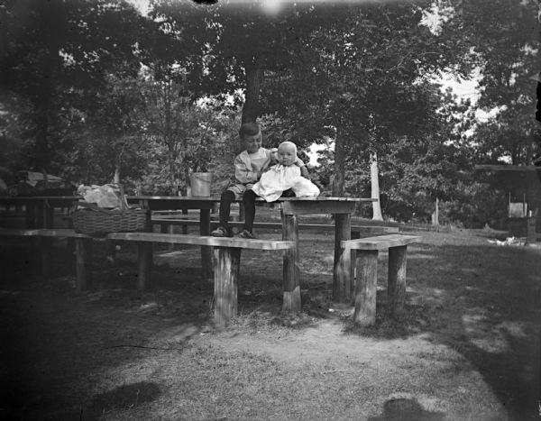 Outdoor portrait of a small boy and infant sitting together on top of a picnic table near a picnic basket.