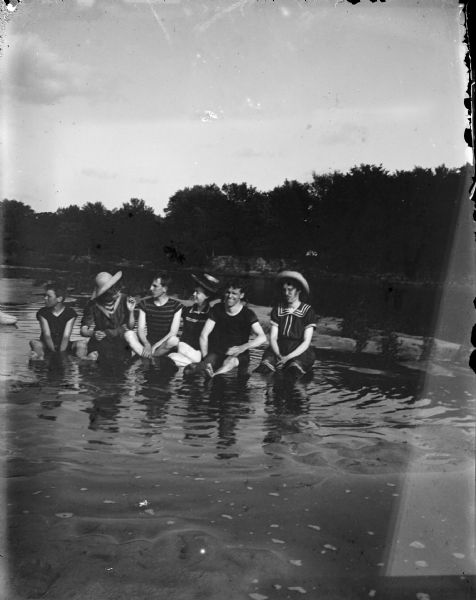 Group portrait of three women, two men, and a boy sitting in the shallow water near a shoreline.
