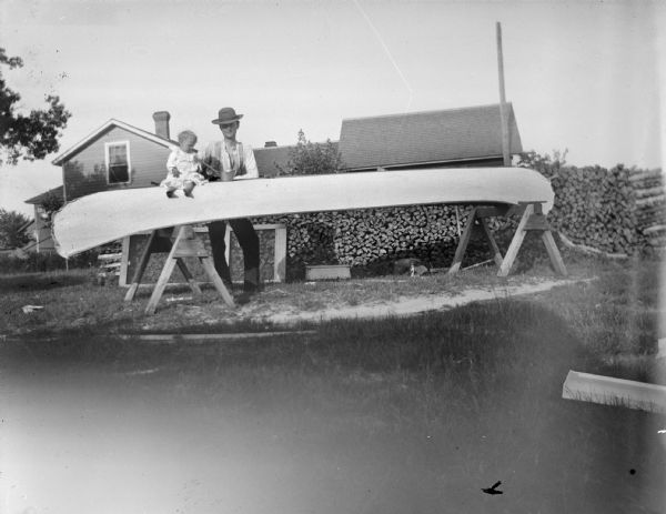 Portrait of a man standing behind a canoe next to a small child sitting on top of it. Behind the canoe is stacked fuelwood and a building.