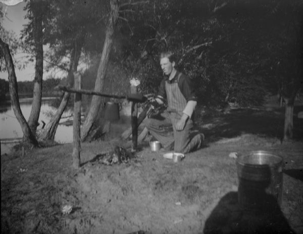 View of a man kneeling by a kettle over a campfire. Identified as camping at Perry Creek.