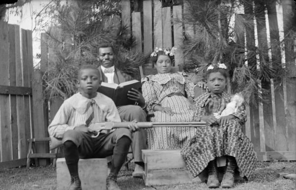 Outdoor group portrait of an African-American family. Sitting in the back is a man, holding a book, and a woman. Sitting in front is a boy, holding a gun, and a girl, holding a doll. The group is sitting in front of a wooden fence.