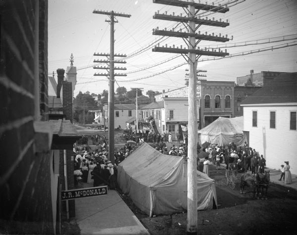 Elevated view of tents and a large group of a people at the intersection of two town streets. Identified as tent shows at the intersection of Main and First Streets in Black River Falls. The steeple of the Catholic church is in the distance on North First Street.