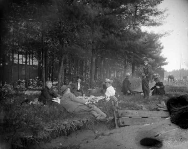 Men and women are sitting and standing near a stand of trees. In the background is a wooden building, and a horse on the right.