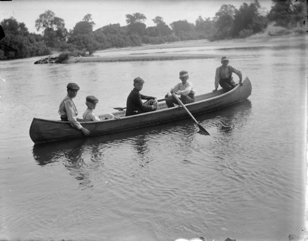 View across water towards five boys sitting in a canoe on the water. The far shoreline is in the background.