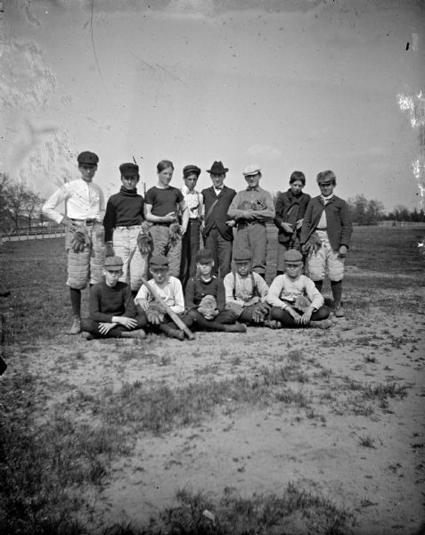 Outdoor group portrait of a group of boys posing sitting and standing in a field. They are wearing uniforms and holding baseball equipment. Identified as the baseball team in Black River Falls, circa 1915 to 1920. The boy in the lower left is identified as Eugene Johnson.