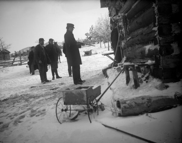 Men standing near a man holding a gun near a log cabin on snow covered ground. Another man is standing in the doorway.