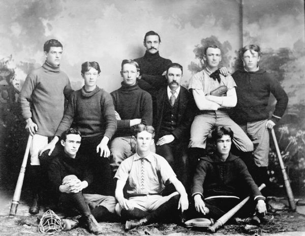 Copy photograph of a studio portrait in front of a painted backdrop of young men standing and sitting. They are wearing uniforms and holding baseball equipment.