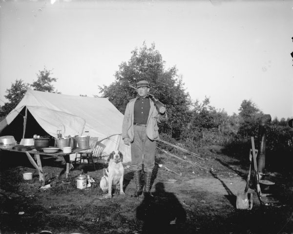 Outdoor portrait of a man holding a rifle standing next to a dog. Behind  them is a campsite with a table and tent.