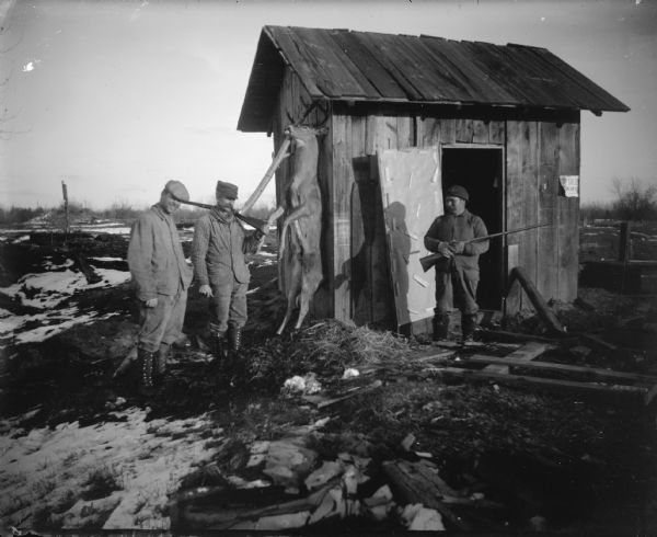 A group of three men are standing next to a wooden hunters' shack with a deer hanging from the roof. Two of the men are holding rifles.
