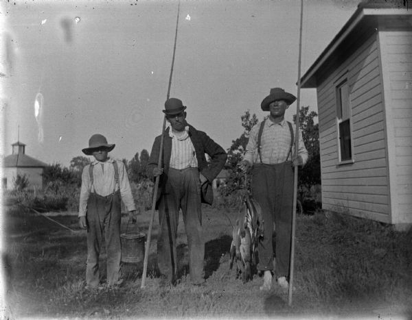 Group portrait of two men and a boy posing standing. The men are holding fishing poles, and the man on the right is holding a stringer of fish.