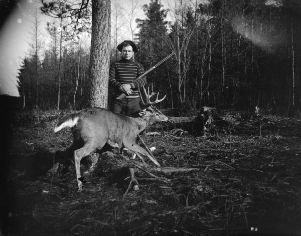 Outdoor portrait of a man posing standing and holding a rifle behind a propped-up dead deer in a forest.