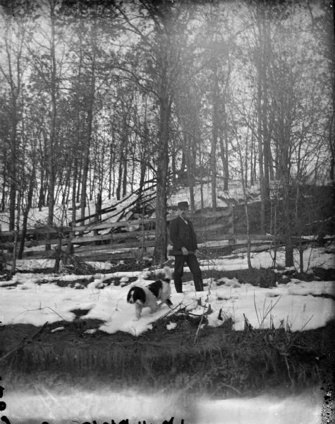 View looking uphill towards a man standing and holding a gun. A dog is nearby on snow-covered ground. Further up the hill are fences.