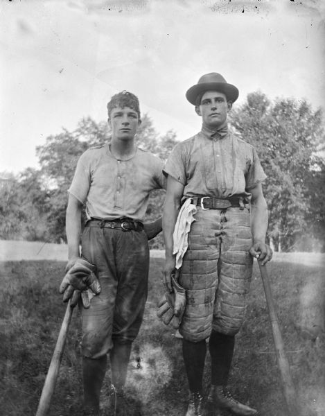 Two men standing outdoors wearing baseball uniforms and holding bats and baseball mitts.