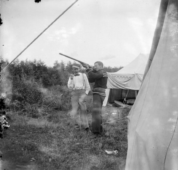 View of two men standing among tents. with one of the men aiming a rifle up into the air.